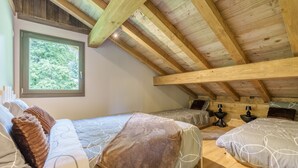 Quarto com teto inclinado, vigas de madeira, uma janela grande e duas camas com roupa de cama estampada.