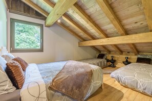 Ein Schlafzimmer mit schrägem Dach, Holzbalken, einem großen Fenster und zwei Betten mit gemusterten Betttextilien.
