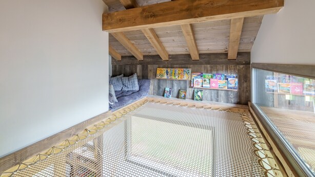 Um quarto com teto inclinado, vigas de madeira, tapete de fibra natural, um sofá e uma parede com livros infantis.