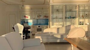 Uma sala de estar moderna com um sofá branco, uma mesa de centro de madeira, e uma área de cozinha com armários e uma mesa de jantar.