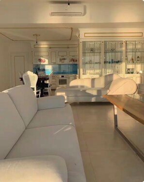 Ein modernes Wohnzimmer mit einem weißen Sofa, einem Holztisch und einer Küchenzeile inklusive Schränken und einem Esstisch.
