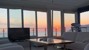 Uma sala de estar moderna com uma televisão grande, sofás brancos, uma mesa de centro de madeira e grandes janelas com vista para o pôr do sol sobre a água.