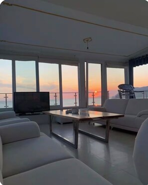 Ein modernes Wohnzimmer mit großem Fernseher, weißen Sofas, einem Holztisch und großen Fenstern mit Blick auf den Sonnenuntergang über dem Wasser.