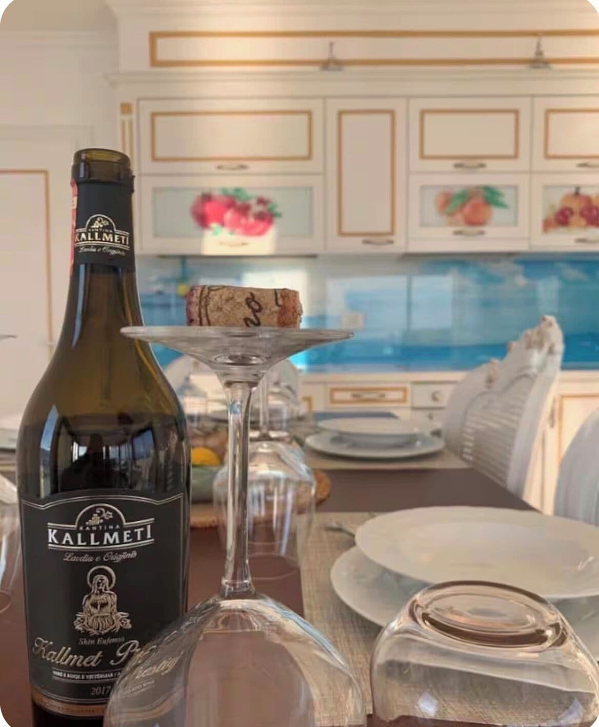 A bottle of Kallmeti wine, a glass with a cork, and a dining table set in a kitchen with white cabinets and a view of the sea.