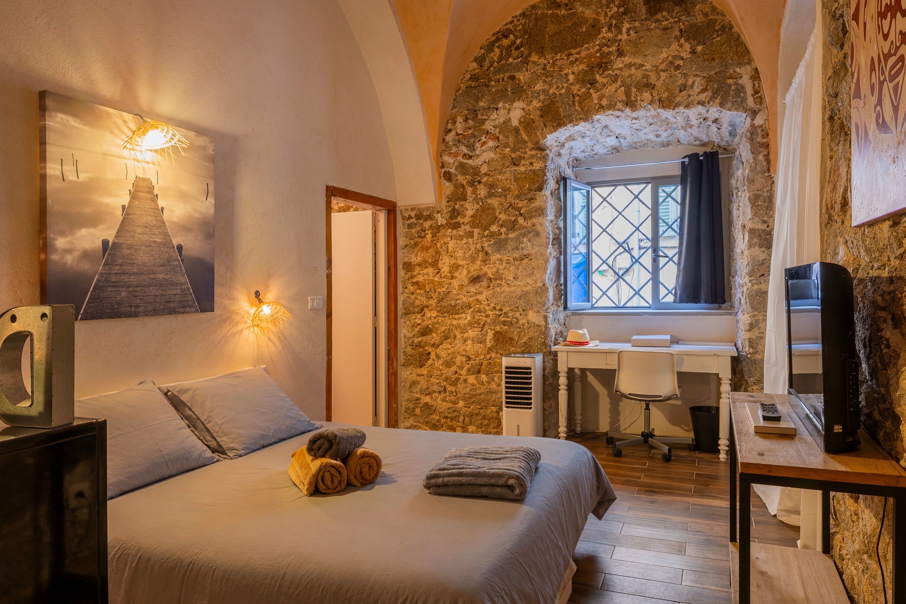A bedroom with a bed, a desk, a chair, a TV, and a stone wall.