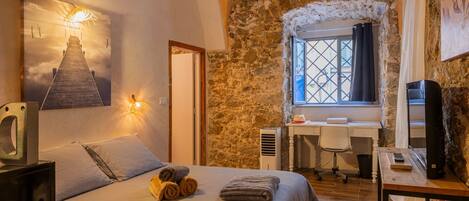 A bedroom with a bed, a desk, a chair, a TV, and a stone wall.