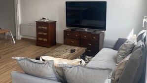 Ein Wohnzimmer mit Flachbildfernseher, Couchtisch aus Holz, grauem Sofa und einer Holzkommode.