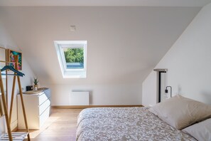Ein Schlafzimmer mit schrägem Dach, einem Oberlicht, einem Bett, einer Kommode und einem kleinen Tisch mit einer Pflanze.