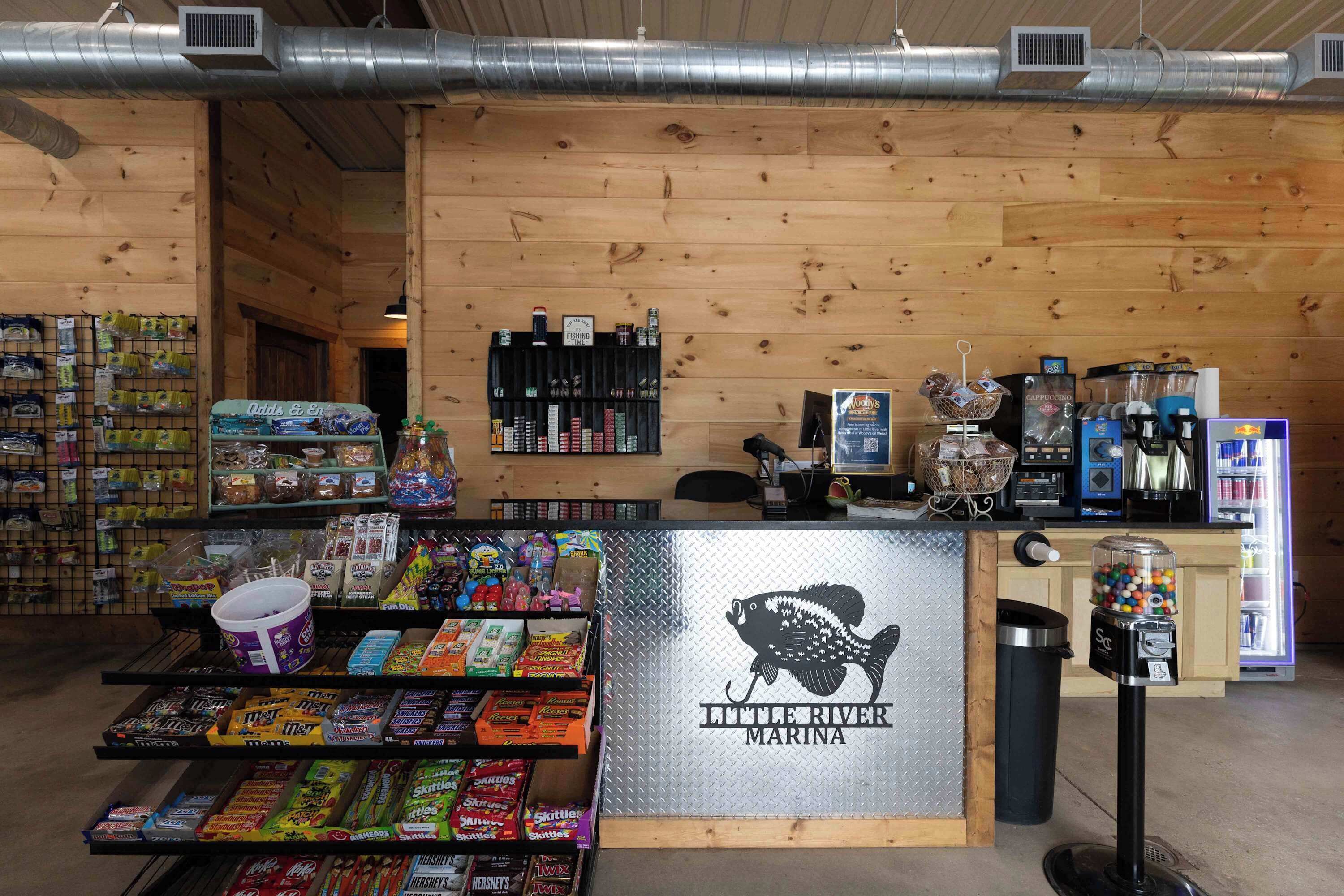 Een kiosk voor snacks en drankjes met diverse lekkernijen, een automaat en een bordje met 'Little River Marina'.