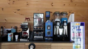 Estação de café com diferentes máquinas de preparo e geladeira abastecida com bebidas.