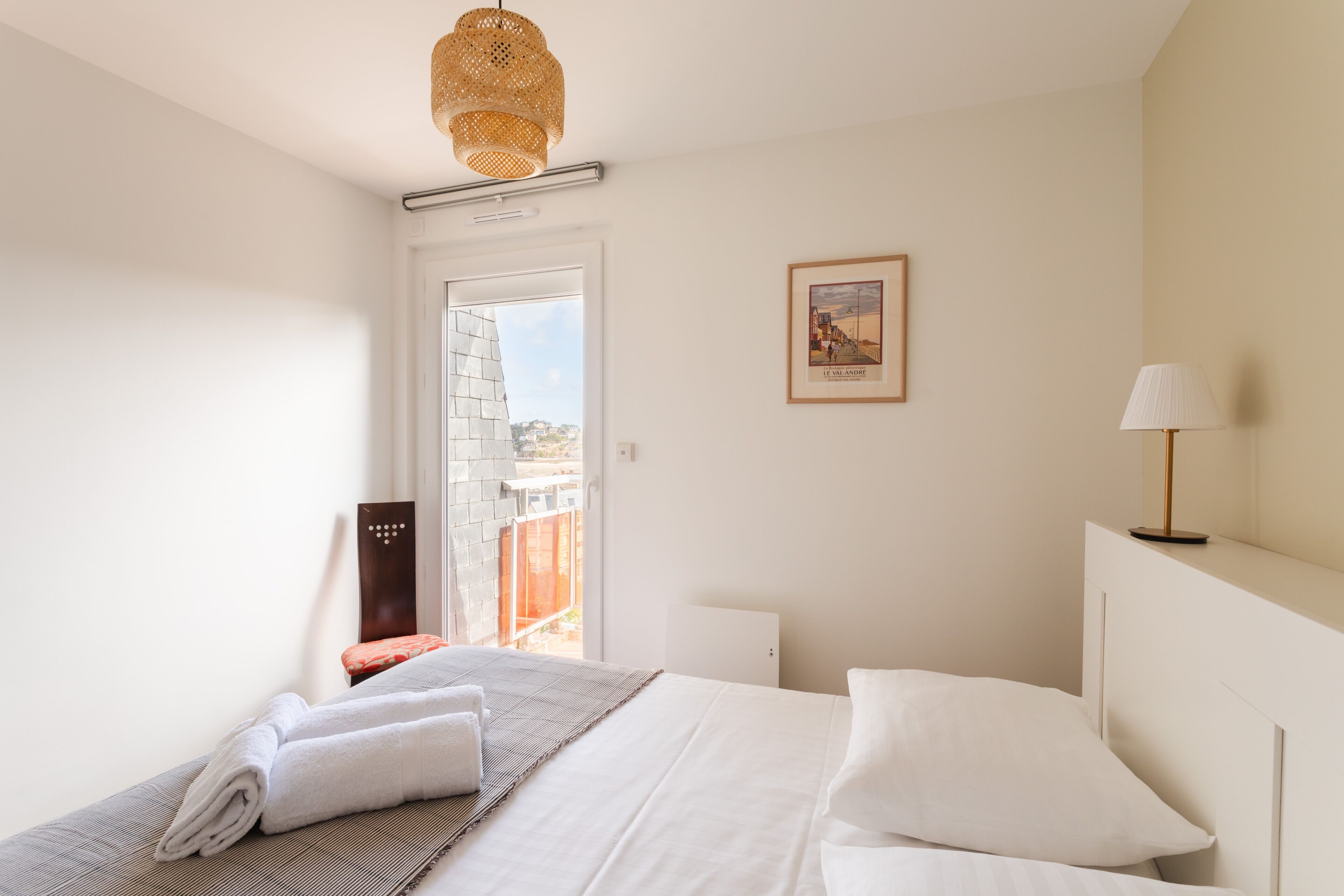 A hotel room with a bed, a window with a view, a framed picture on the wall, and a lamp.