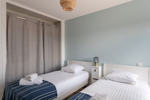 A hotel room with two single beds, a bedside table with a lamp, and a window with curtains. - Hirta - À 100 m de la plage et vue mer (Pléneuf-Val-André)