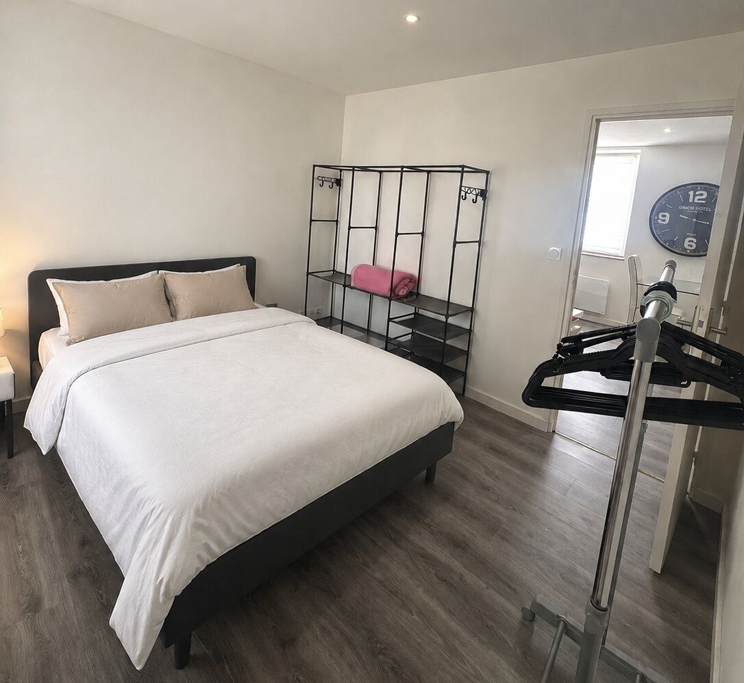 A hotel room with a bed, a metal rack with clothes, and a clock on the wall.
