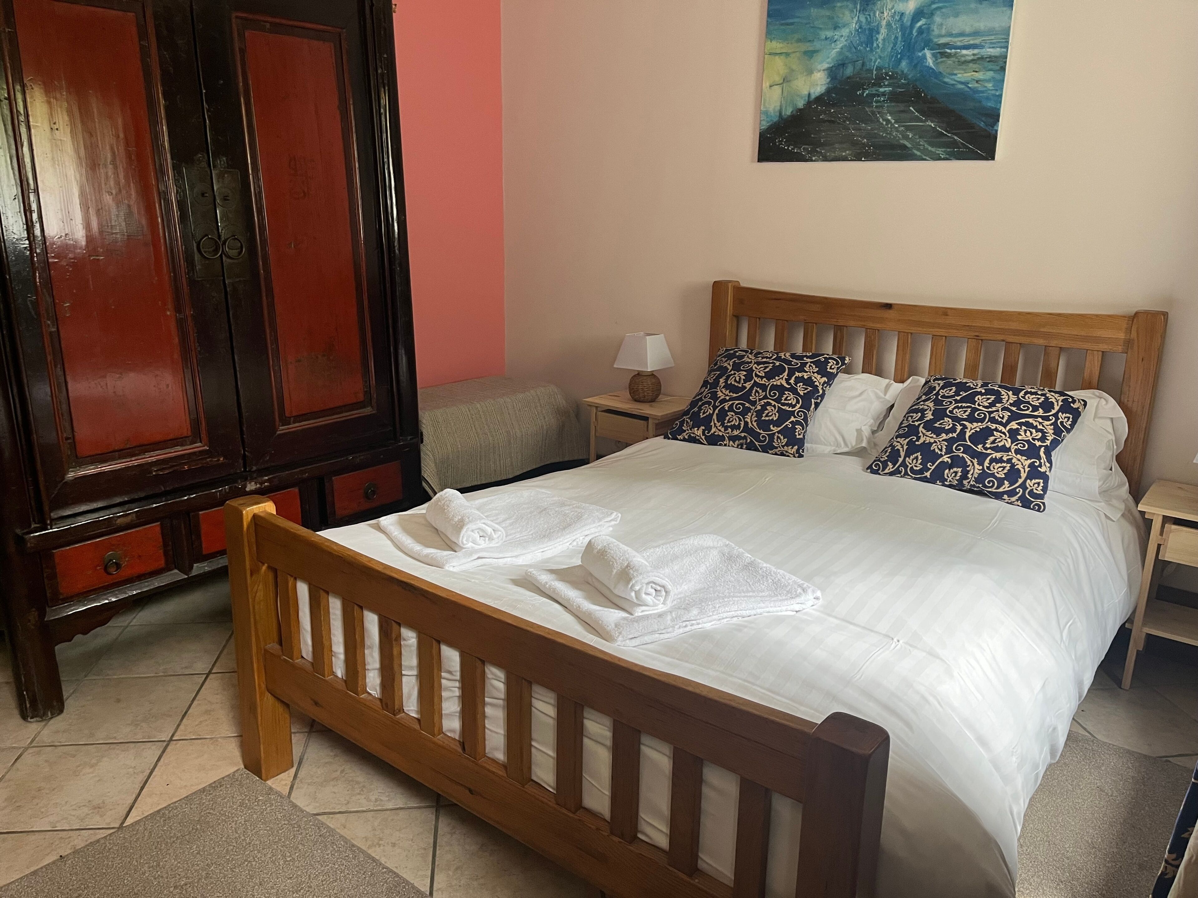 Una cama bien hecha con sábanas blancas y toallas dobladas, un cabecero de madera, una mesita de noche con una lámpara y un cuadro en la pared.