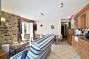 A cozy room with a stone wall, a dining table with chairs, a striped sofa, and wooden cabinets. - Degas - Appt pour 6 avec terrasse et parking (Dinan)