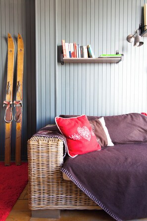 Una habitación con un sofá de mimbre, un cojín rojo con estampado de corazón y un par de esquíes apoyados en la pared.