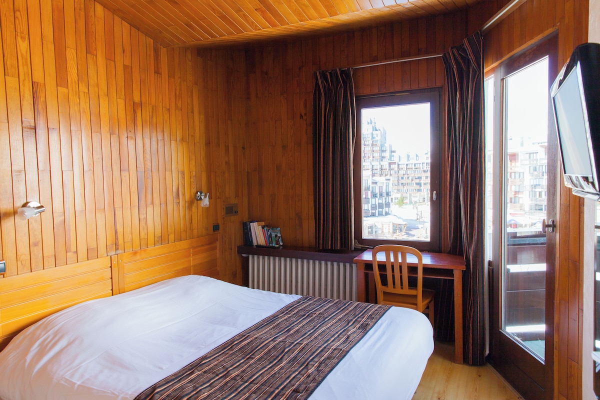 A hotel room with a wooden paneled wall, a bed with a white mattress, a desk with a chair, a radiator, and a window with curtains.