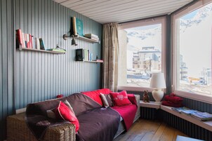 A cozy room with a wooden floor, a red sofa with cushions, a bookshelf, a window with a view of buildings, and a lamp on a side table.