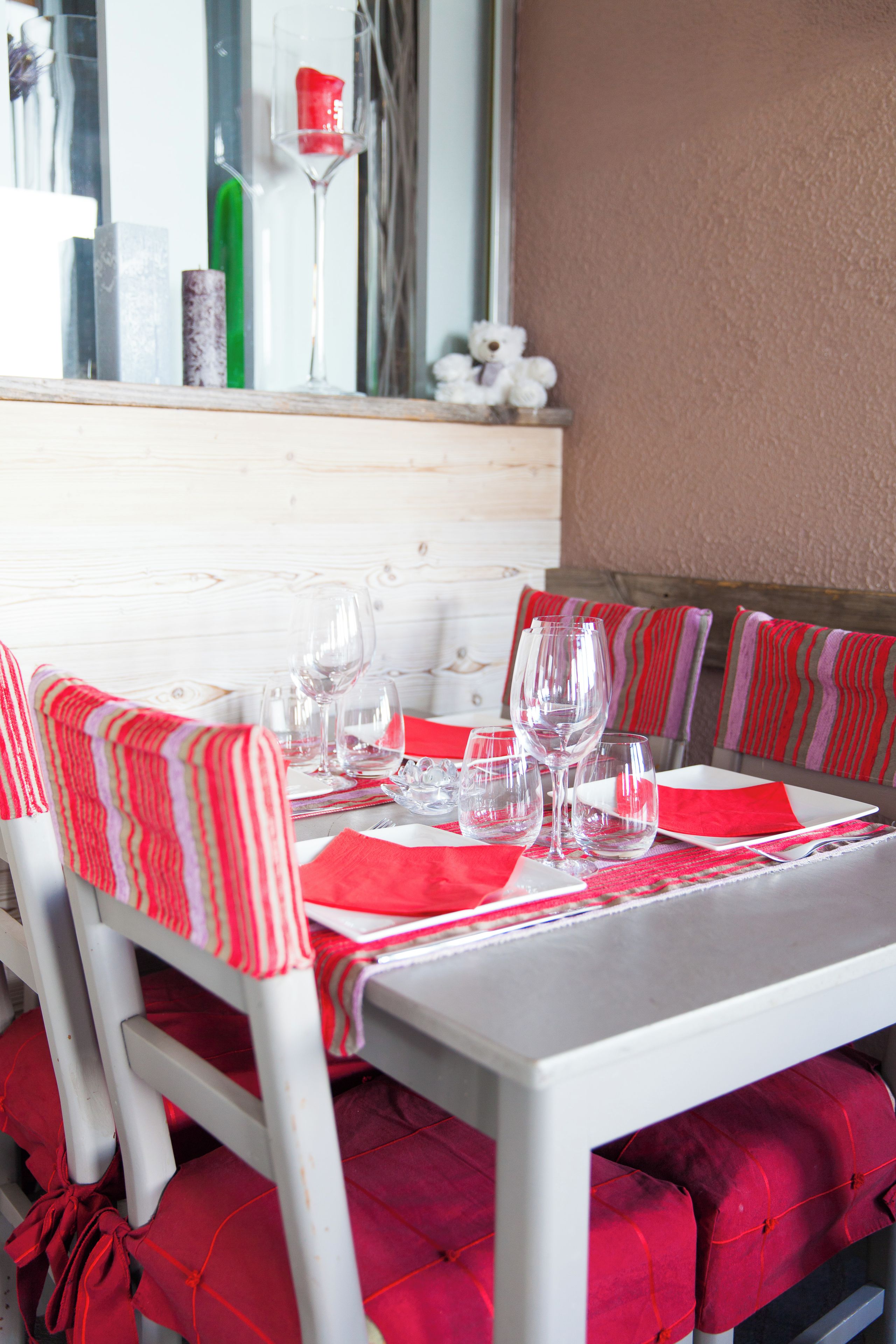 Une table à manger dressée avec des verres à vin, des assiettes et des couverts, entourée de chaises dotées de coussins rouges.