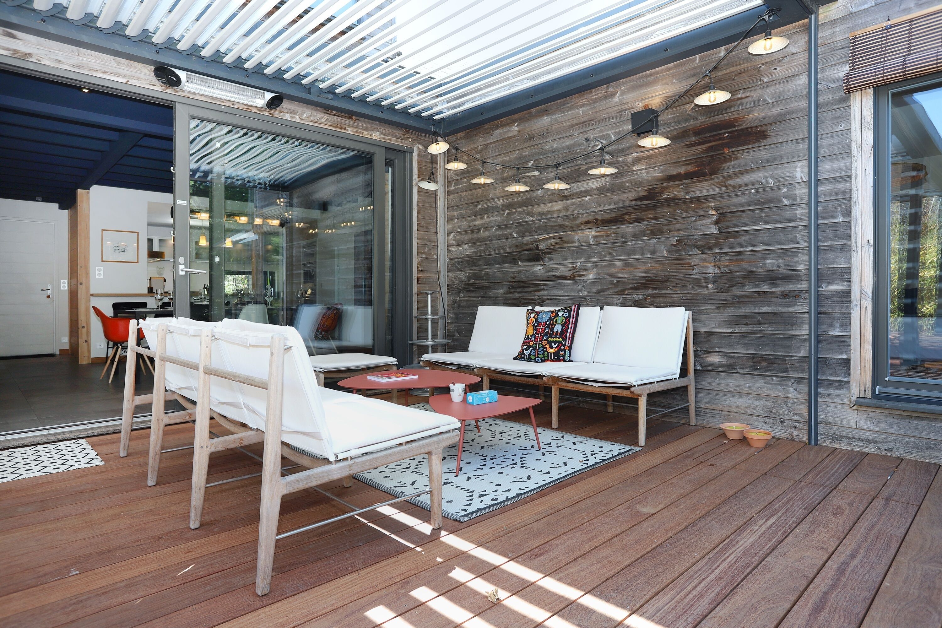 Un espacio de esparcimiento moderno al aire libre con puertas de vidrio que dan a un ambiente interior, piso de madera y vistas del exterior.