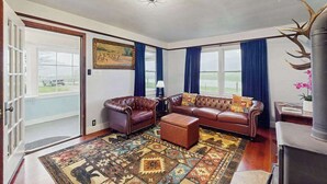 Ein Wohnzimmer mit einer braunen Ledersofa-Garnitur, einem gemusterten Teppich und einem großen Fenster mit blauen Vorhängen.