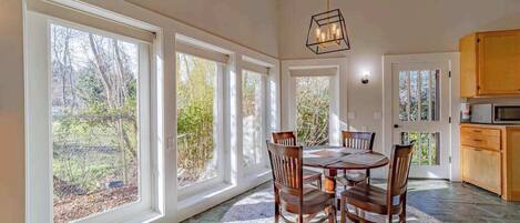 A dining area with a round table and chairs, large windows, and a kitchen area with wooden cabinets.