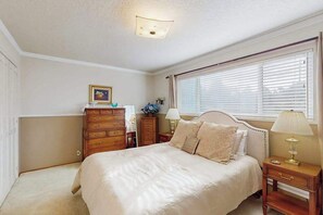 A bedroom with a bed, dresser, nightstand, lamp, and window with blinds. - West End Wanderlust - Stylish Coastal Escape (Eureka)