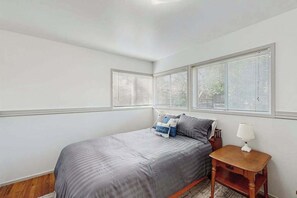A bedroom with a bed, a nightstand, and a window with blinds. - West End Wanderlust - Stylish Coastal Escape (Eureka)