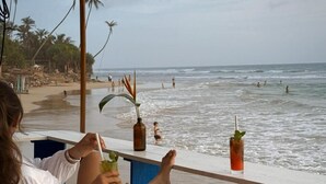 Uma pessoa sentada perto de um banco de madeira, com vista para uma praia com palmeiras e o oceano.