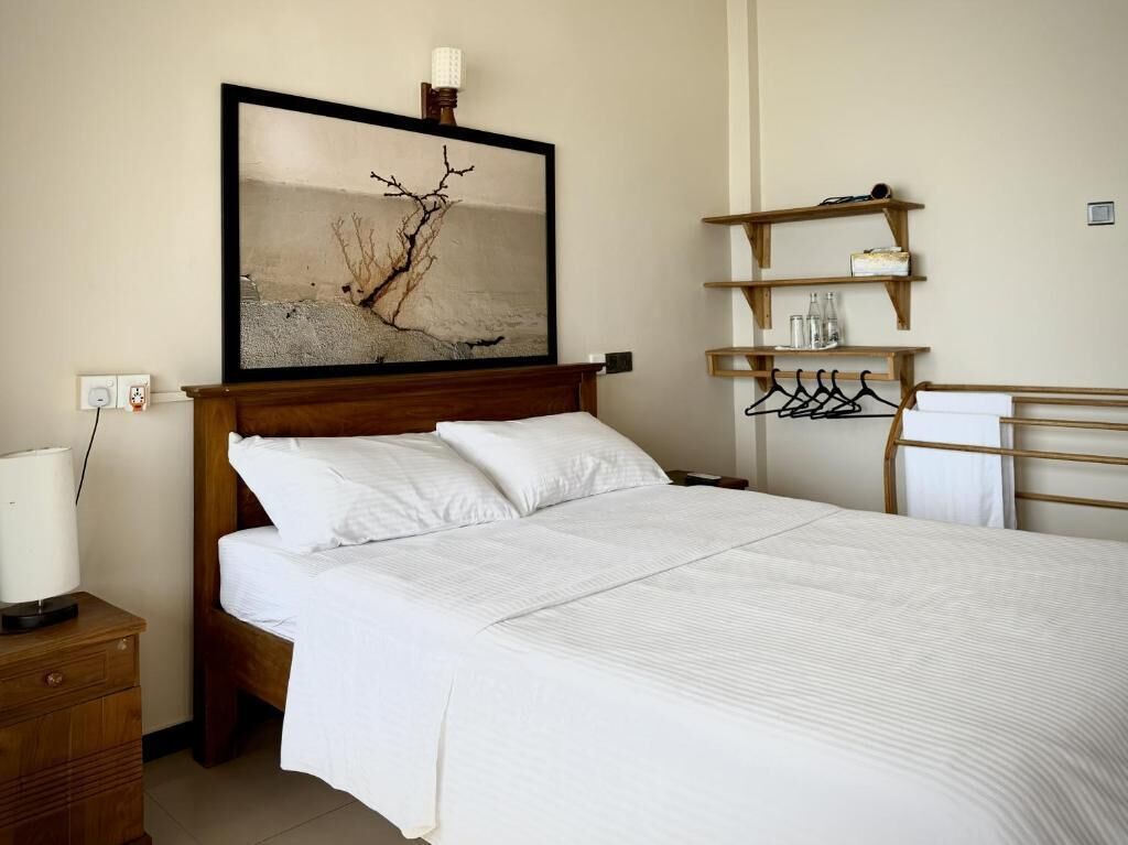 Un lit bien fait, avec du linge de lit blanc, une tête de lit en bois, un cadre photo fixé au mur et une étagère en bois avec des cintres et des bouteilles.