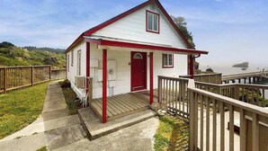 A small white house with a red door and porch, located near a body of water with a wooden dock. - Seascape Home - Spacious Coastal Retreat with Ocean Views (Trinidad)