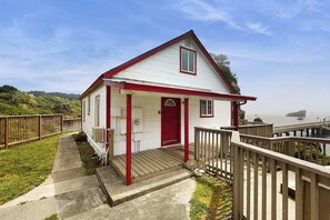 Ein kleines weißes Haus mit roter Tür und Veranda, in der Nähe eines Gewässers mit Holzsteg.