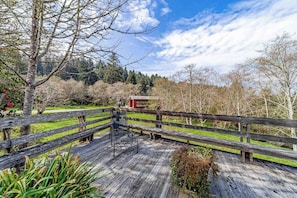 Eine Holzterrasse mit einer Bank und einem Tisch, umgeben von einem Holzzaun, mit Blick auf eine grüne Landschaft mit Bäumen und einem Gebäude in der Ferne.