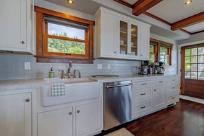 A kitchen with white cabinets, a stainless steel dishwasher, and a window with a view of greenery. - Trinidad Pearl - Oceanfront, Hot Tub, Pickleball (Trinidad)