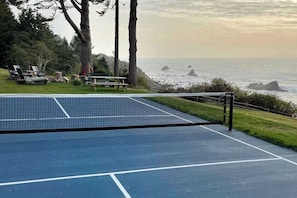 A tennis court with a net, surrounded by chairs and a picnic table, overlooking a coastal area with rocks and trees. - Trinidad Pearl - Oceanfront, Hot Tub, Pickleball (Trinidad)