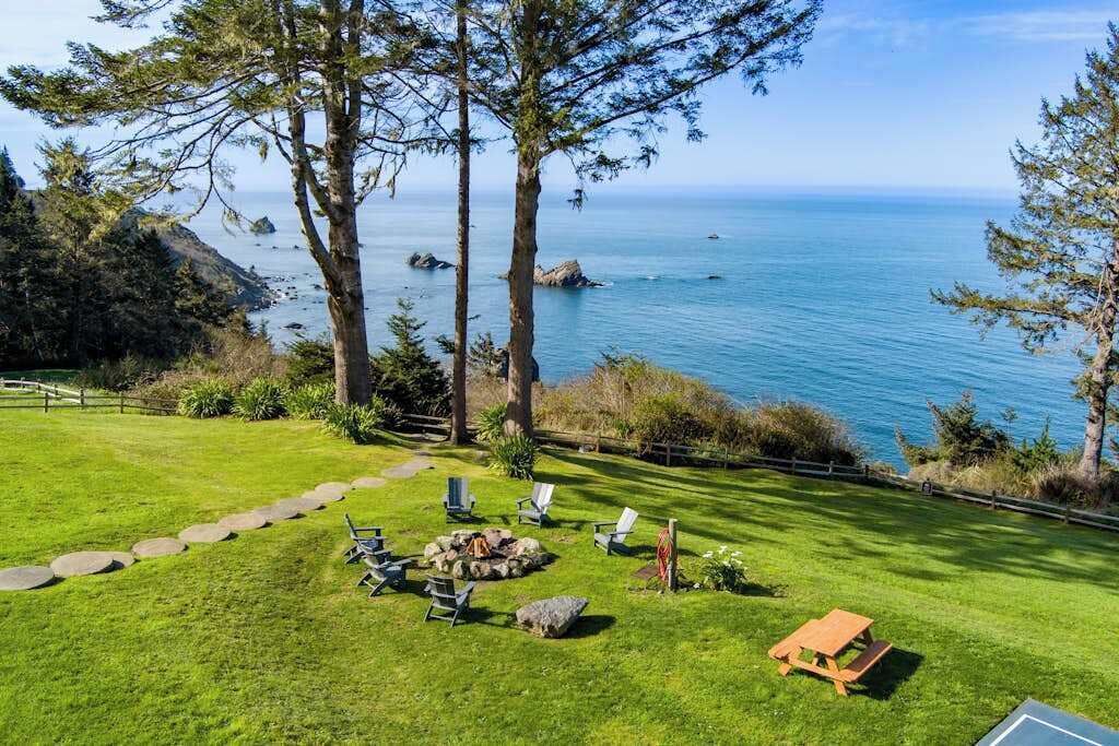 A grassy outdoor area with a wooden deck, lounge chairs, and a picnic table overlooking a body of water.