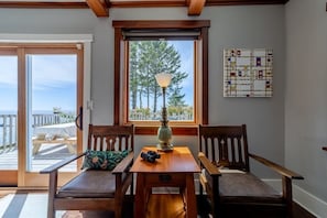 A room with a wooden table and chairs, a lamp, and a window with a view of trees and a deck. - Trinidad Pearl - Oceanfront, Hot Tub, Pickleball (Trinidad)