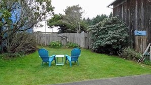 A grassy outdoor area with a wooden fence, a blue chair, a small table, and a blue tarp on the fence. - Ocean Treasures - Charming Coastal Home Near the Beach (Smith River)
