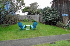 A grassy outdoor area with a wooden fence, a blue chair, a small table, and a blue tarp on the fence. - Ocean Treasures - Charming Coastal Home Near the Beach (Smith River)