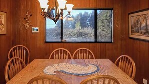 A dining area with a wooden table and chairs, a chandelier, and a window with a view of trees. - Ocean Treasures - Charming Coastal Home Near the Beach (Smith River)
