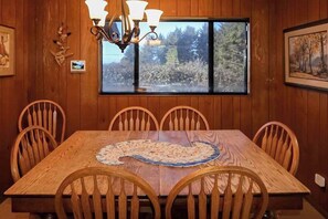 A dining area with a wooden table and chairs, a chandelier, and a window with a view of trees. - Ocean Treasures - Charming Coastal Home Near the Beach (Smith River)