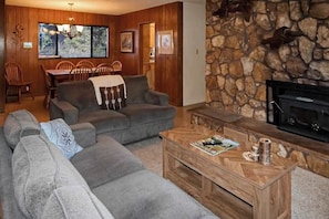 A living room with a stone wall, a fireplace, a sofa, a coffee table, and a dining area with wooden furniture. - Ocean Treasures - Charming Coastal Home Near the Beach (Smith River)