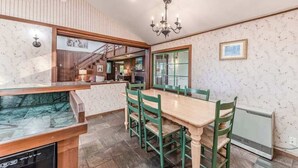 Een eetgedeelte met een houten tafel en groene stoelen, een kroonluchter, en uitzicht op een keuken en een trap.