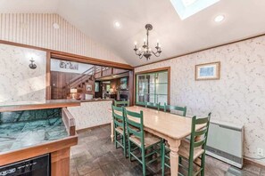 A dining area with a wooden table and green chairs, a chandelier, and a view of a kitchen and staircase. - Seaside Meadow - Peaceful Coastal Retreat Surrounded by Nature (Trinidad)