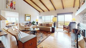 Ein geräumiges Wohnzimmer mit hoher Decke, Holzbalken, einem großen Fenster und vielfältigen Sitzgelegenheiten.