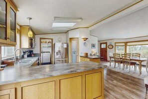 A kitchen with wooden cabinets, a built-in island, a dining area with a table and chairs, and a view through the window. - Pacific View Escape - Bay Views & Jetted Tub (Loleta)
