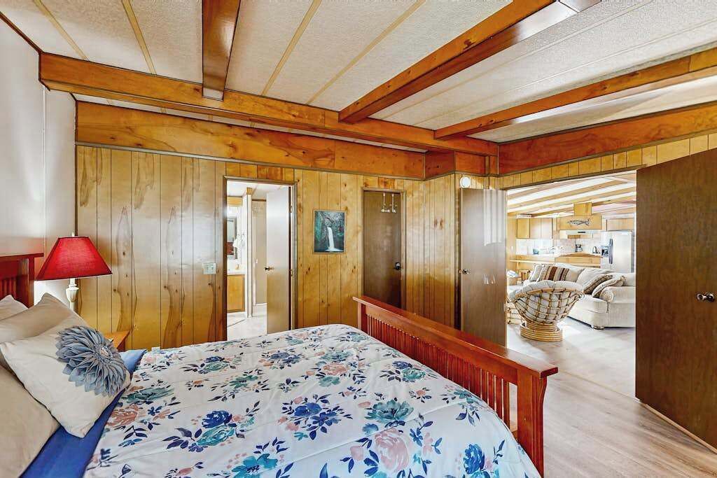 A bedroom with wooden paneling, a bed with a floral bedspread, a red lamp, and a view into a living area with a sofa and a chair.