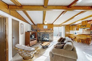 A cozy living room with a stone fireplace, wooden beams, a sofa, and a wicker chair. - Redwood Coast River House (Crescent City)