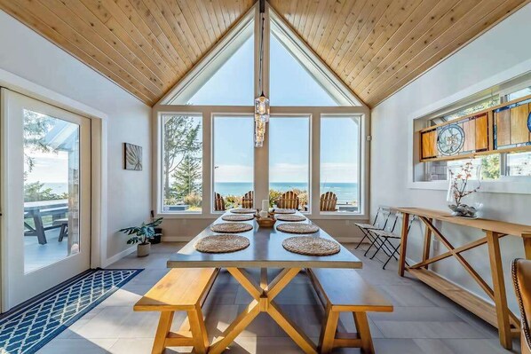 A dining area with a long table, benches, and a view of the sea through large windows. - Seagull's Bluff - Scenic Coastal Retreat with Ocean Views (Trinidad)