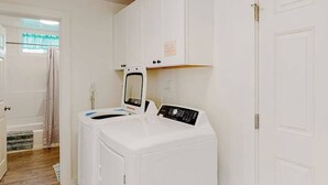 A laundry room with a front-loading washer and dryer, a window with a curtain, and a door leading to another room. - Cedar Street Cottage (Eureka)